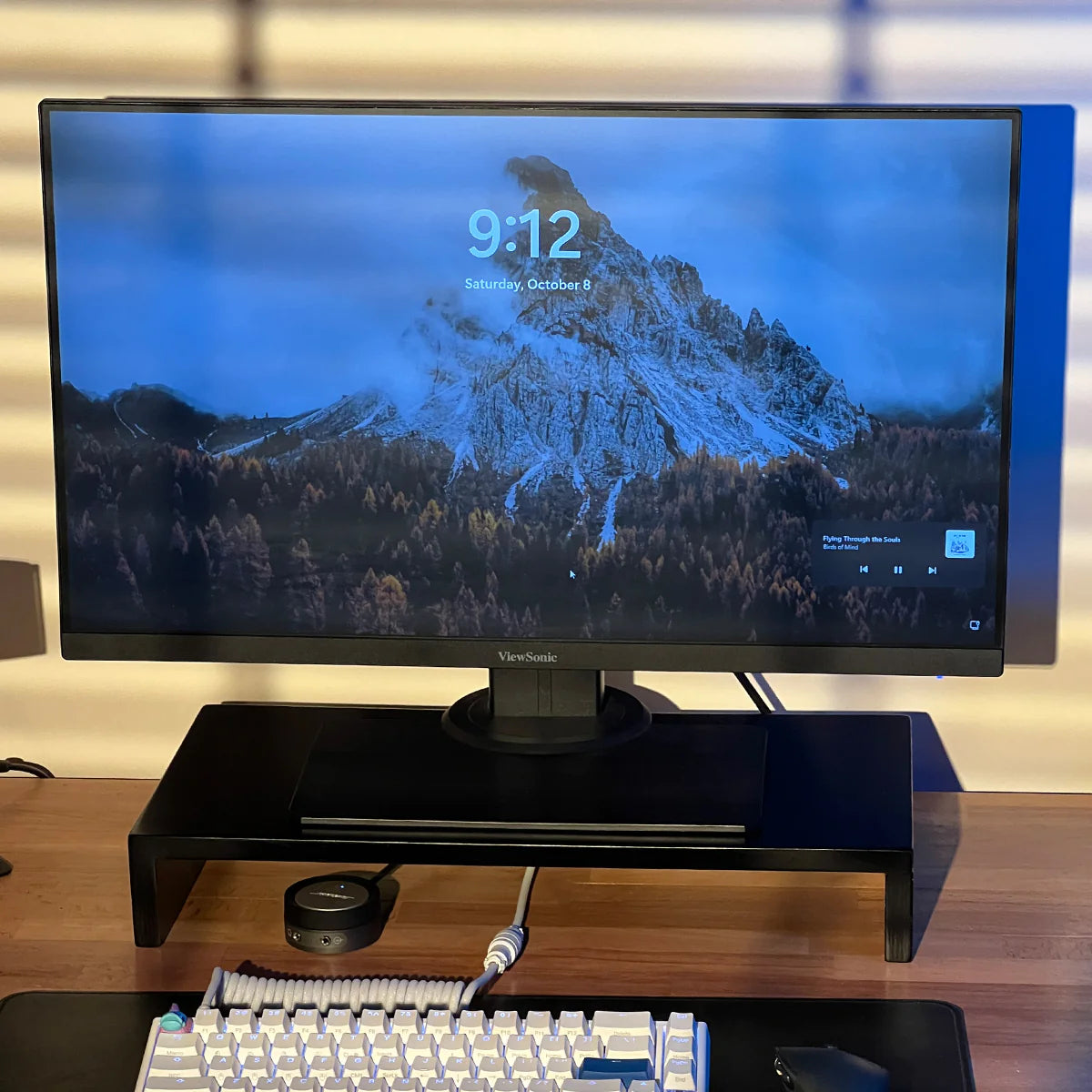 Modern stand riser by woodsy with monitor on desk displaying mountain wallpaper with media player controls, keyboard in foreground.