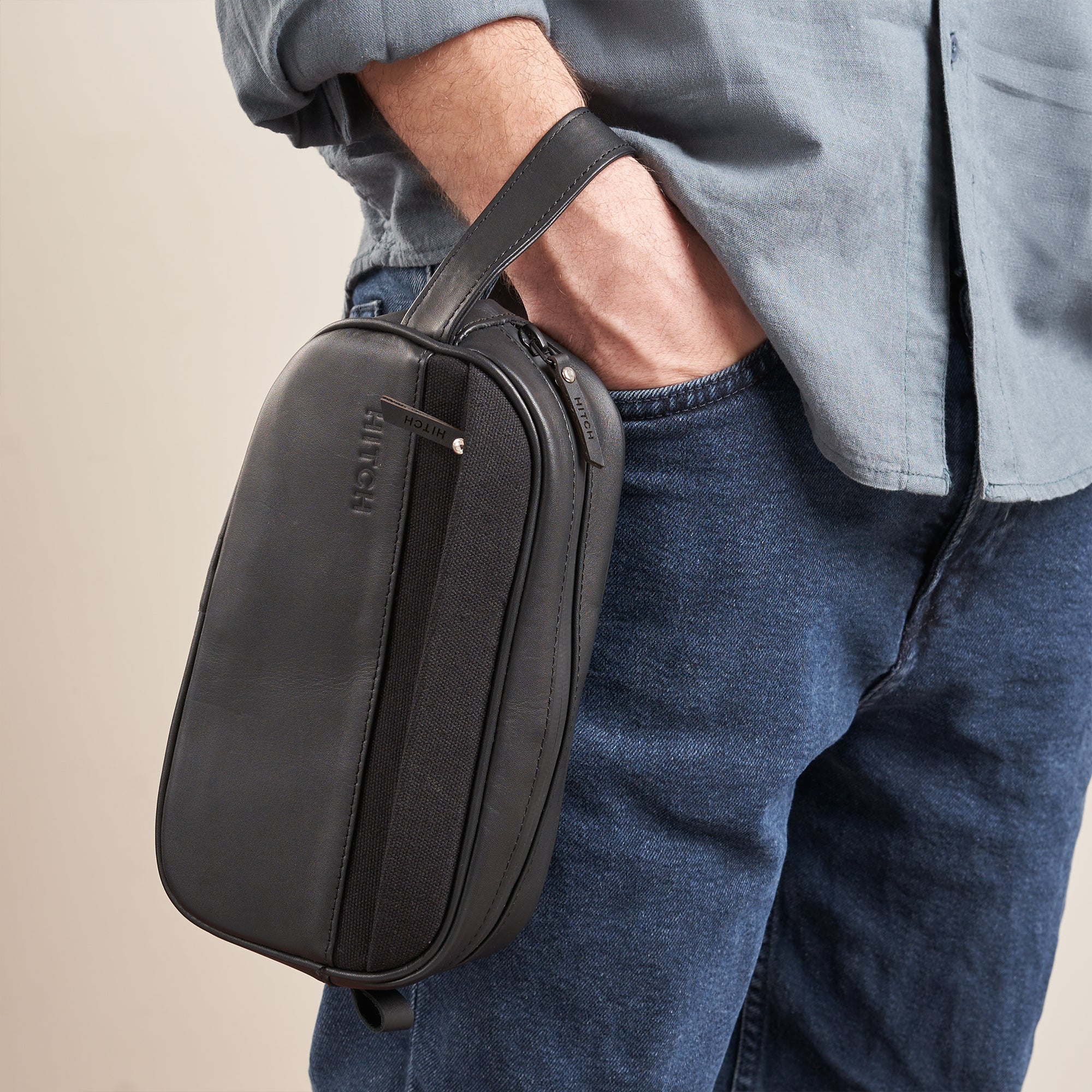 Man carrying a black leather Dopp kit with brand logo HITCH embossed, paired with casual denim and shirt.