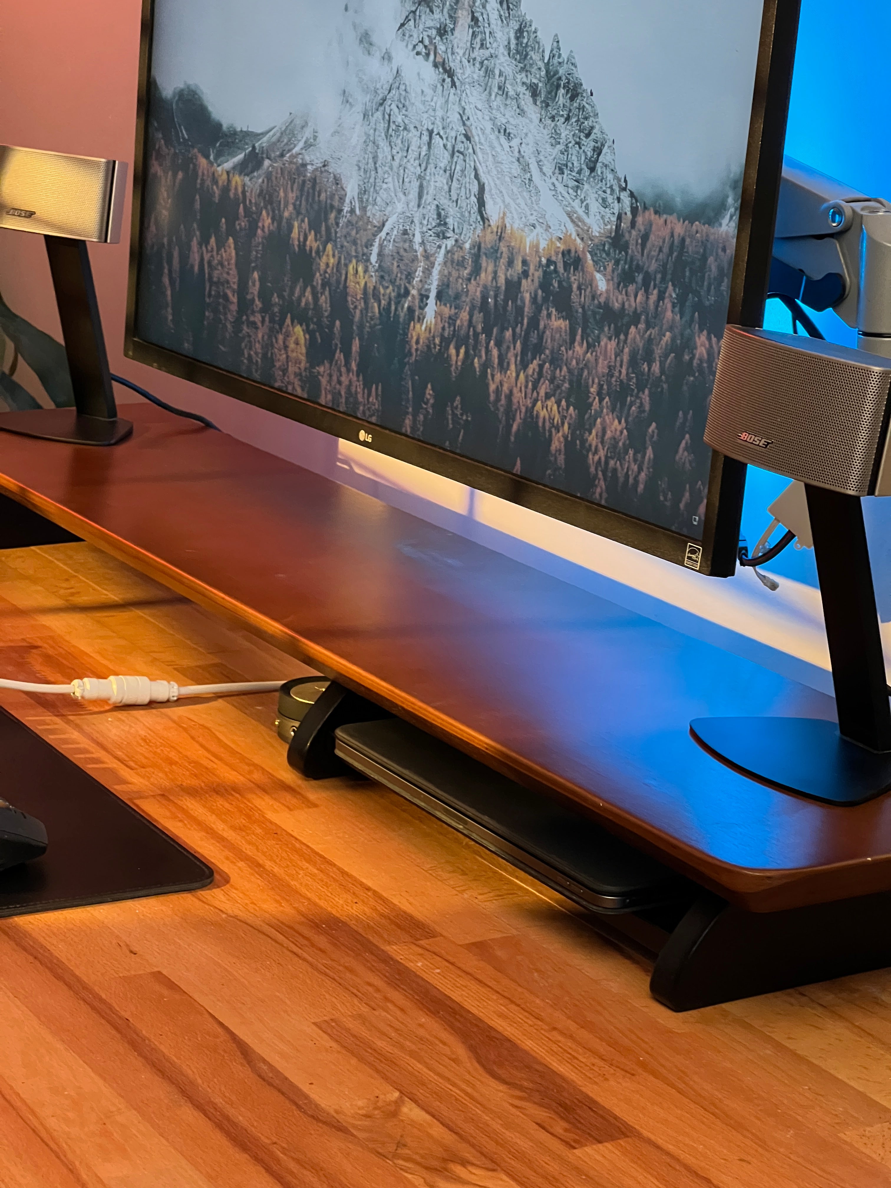 Dual monitors on woodsy wooden desk, sound bar, Bose speaker, and ambient lighting.