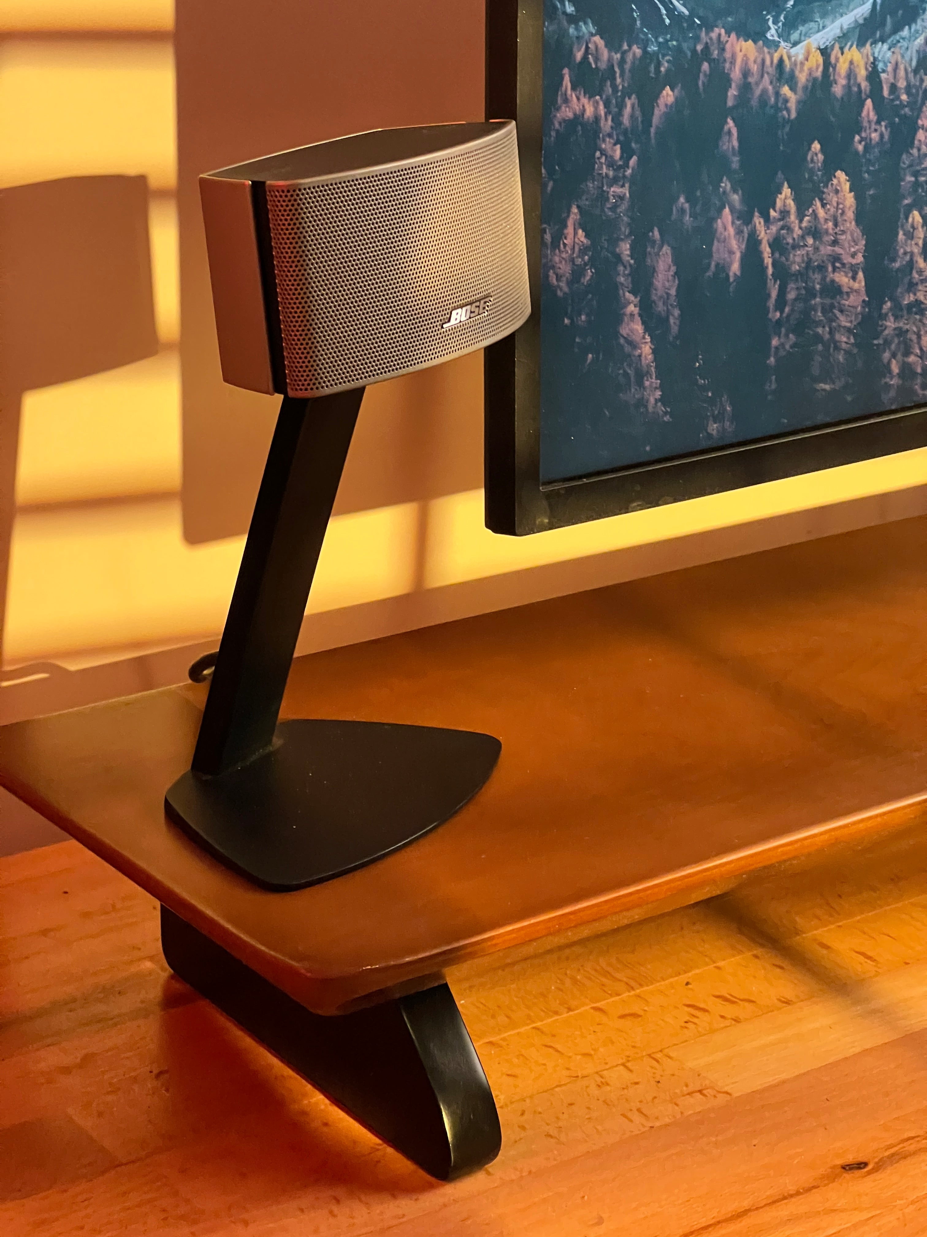 Bose computer speaker on woodsy wooden desk shelf with monitor displaying forest wallpaper.